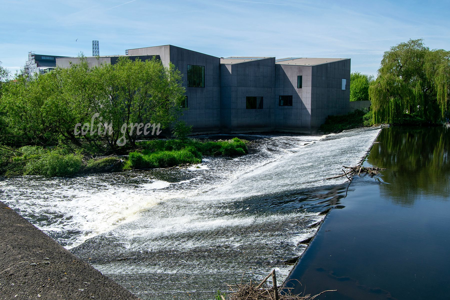 In the foreground, there is a grey building with a flat roof. The building is part of the Hepworth Wakefield, a gallery in Wakefield, England.   * To the left of the building is a body of water, the River Calder,  with a waterfall cascading over rock