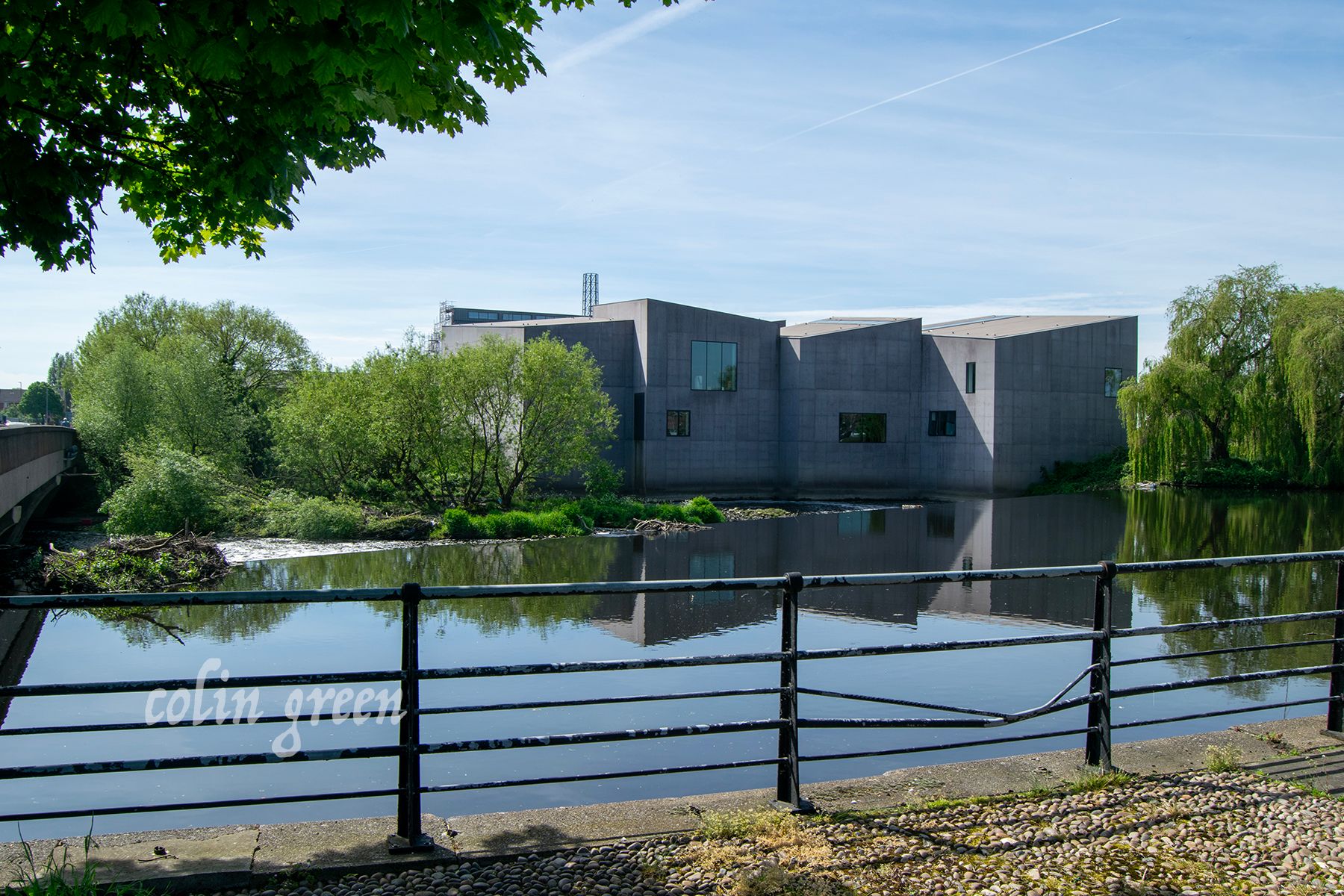 In the foreground, there is a grey building with a flat roof. The building is part of the Hepworth Wakefield, a gallery in Wakefield, England.   * To the left of the building is a body of water, the River Calder,  with a waterfall cascading over rock