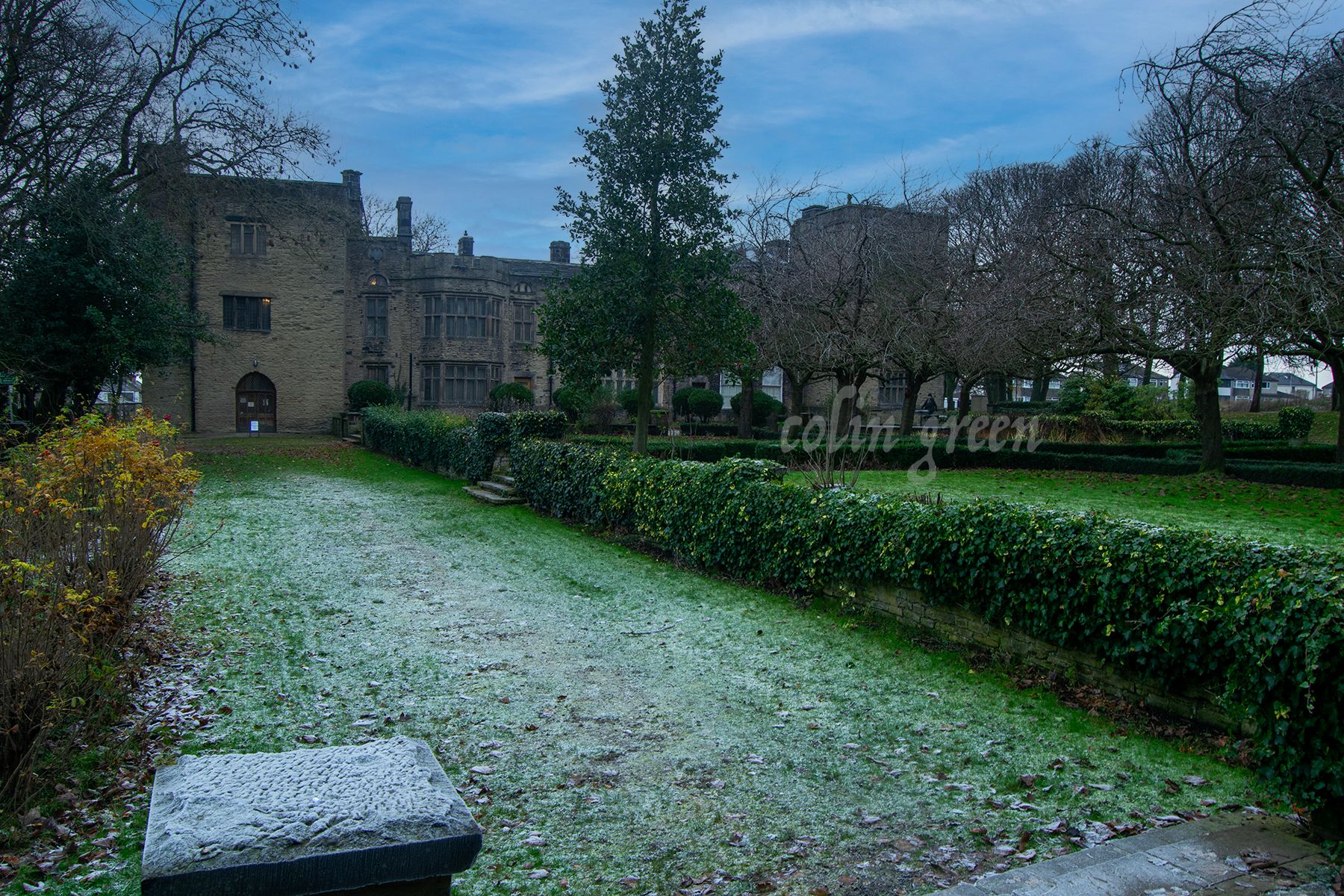 Bolling Hall, stands proudly amidst meticulously manicured lawns. Tall, mature trees provide shade and create a serene atmosphere around the historic building. The hall's stone facade and mullioned windows are complemented by the lush green surroundi