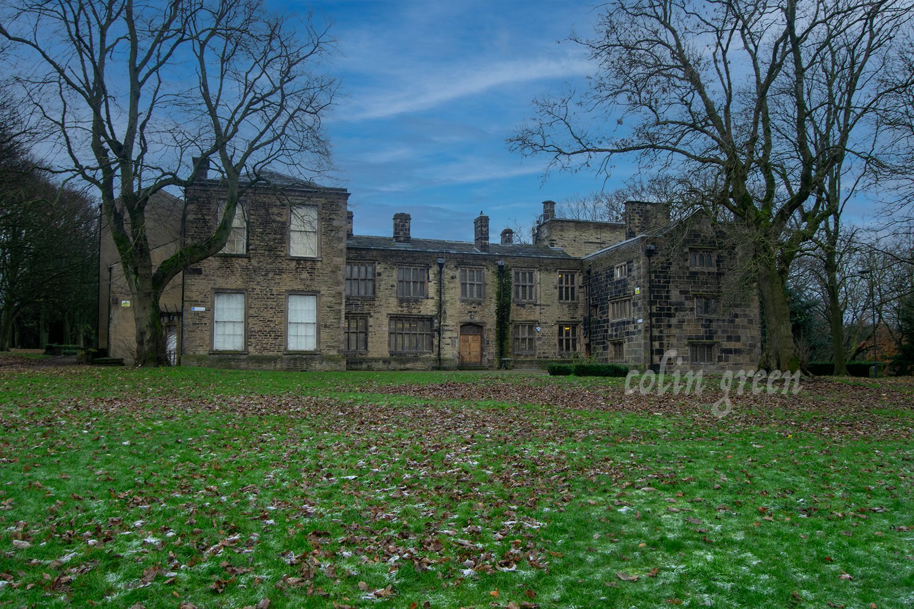 Bolling Hall, stands proudly amidst meticulously manicured lawns. Tall, mature trees provide shade and create a serene atmosphere around the historic building. The hall's stone facade and mullioned windows are complemented by the lush green surroundi