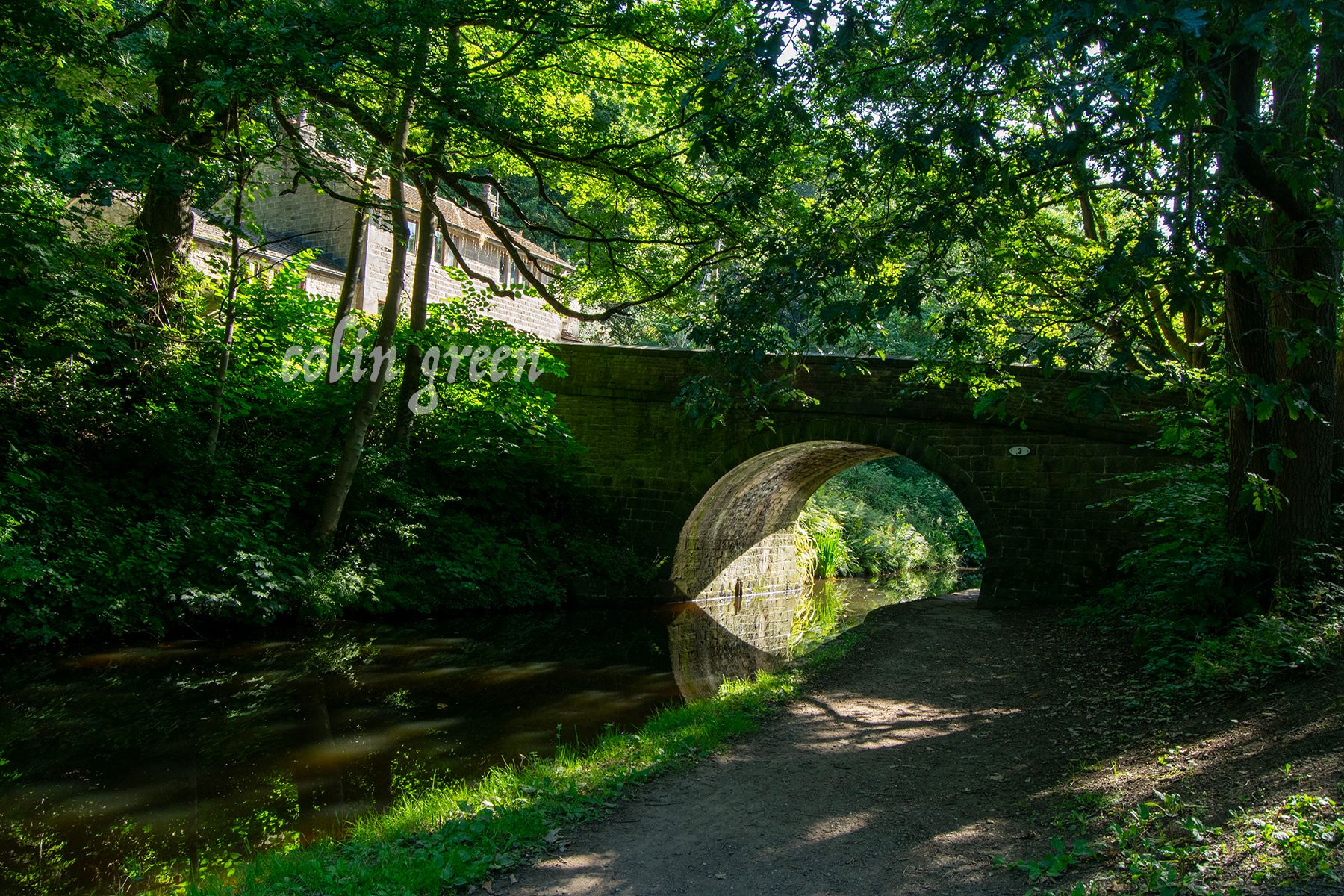 A stone arched bridge over a canal, surrounded by lush greenery and with a path leading under it.