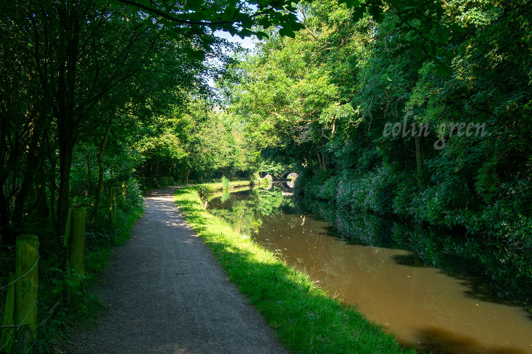 A path runs alongside a canal, shaded by trees. A stone bridge can be seen in the distance.