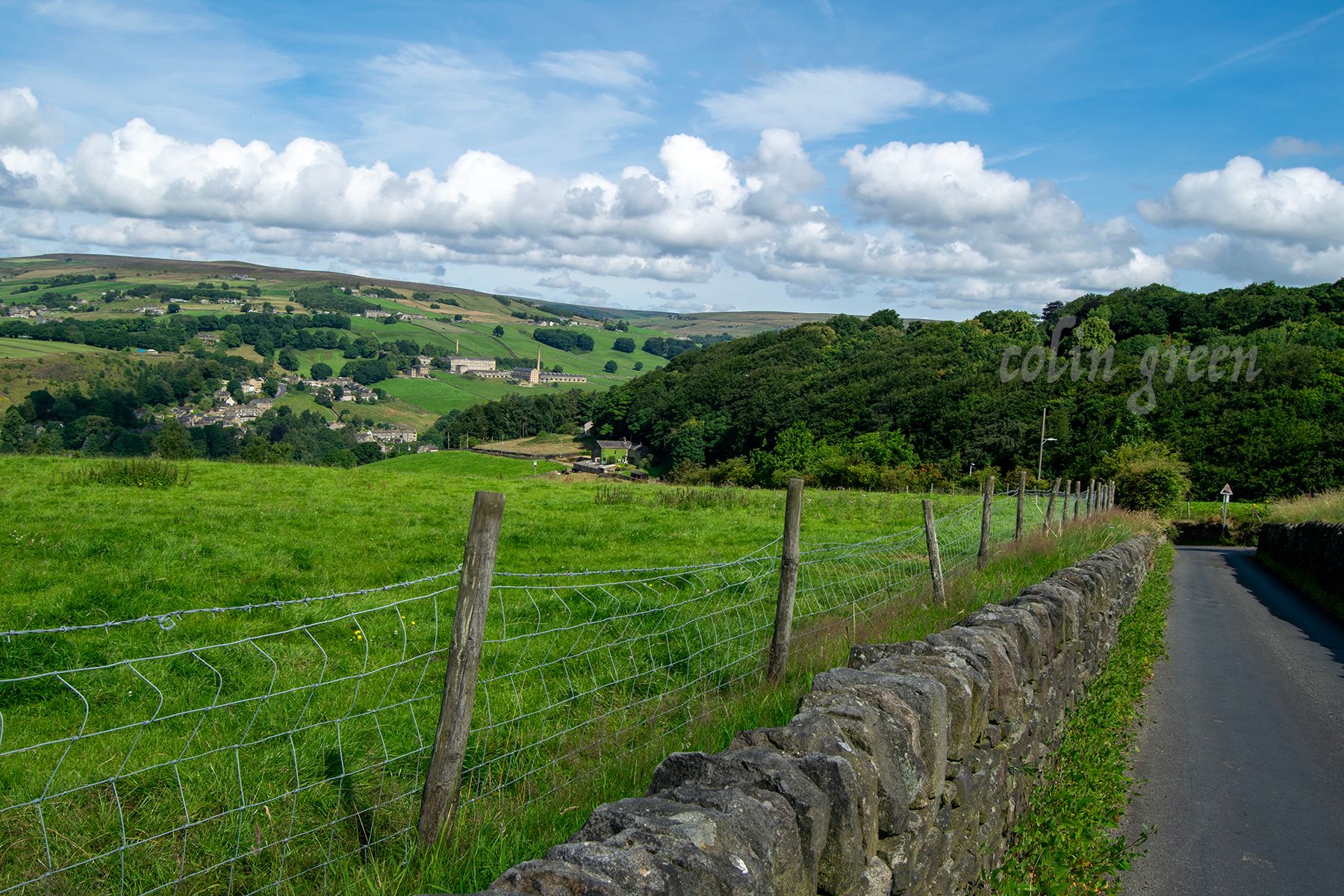 A scenic rural landscape with rolling hills, a small town in the distance, a paved road, a stone wall, and a wire fence. The sky is blue with fluffy white clouds.