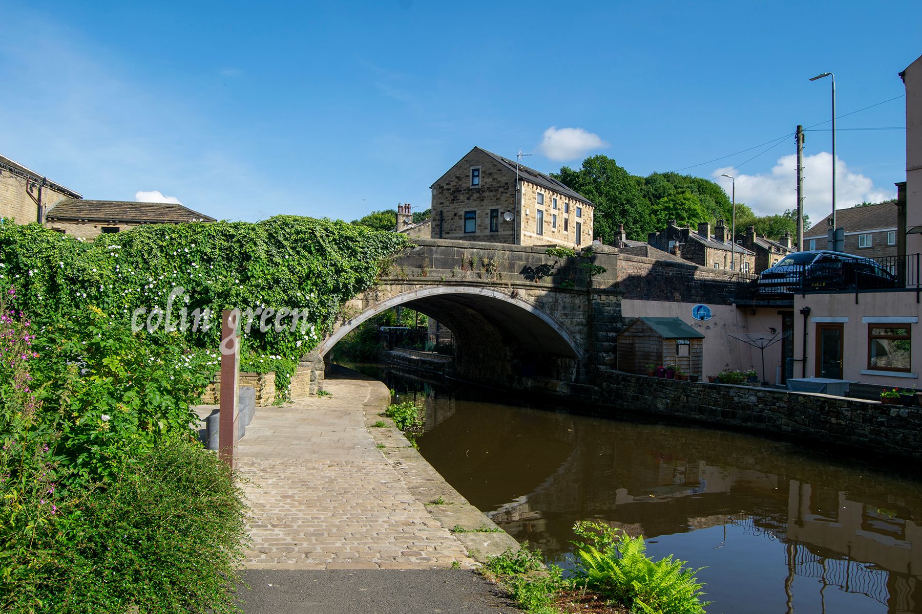 A stone bridge arches over a canal, with lush greenery on either side. A quaint building with a slate roof stands behind the bridge. The water reflects the blue sky and the surrounding buildings.