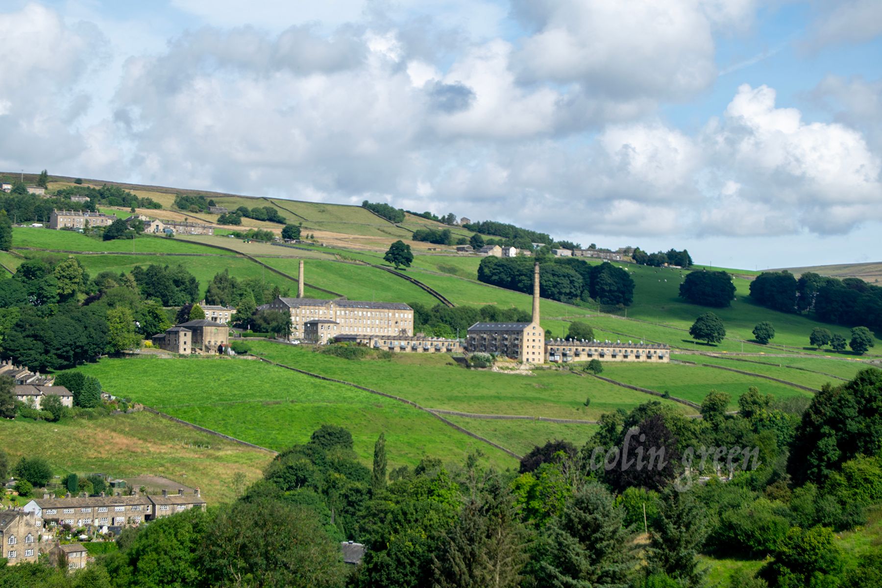 A historic mill complex sits on a hillside, surrounded by rolling green fields and trees. The mill buildings are made of stone and have tall chimneys. The sky is blue with white clouds.