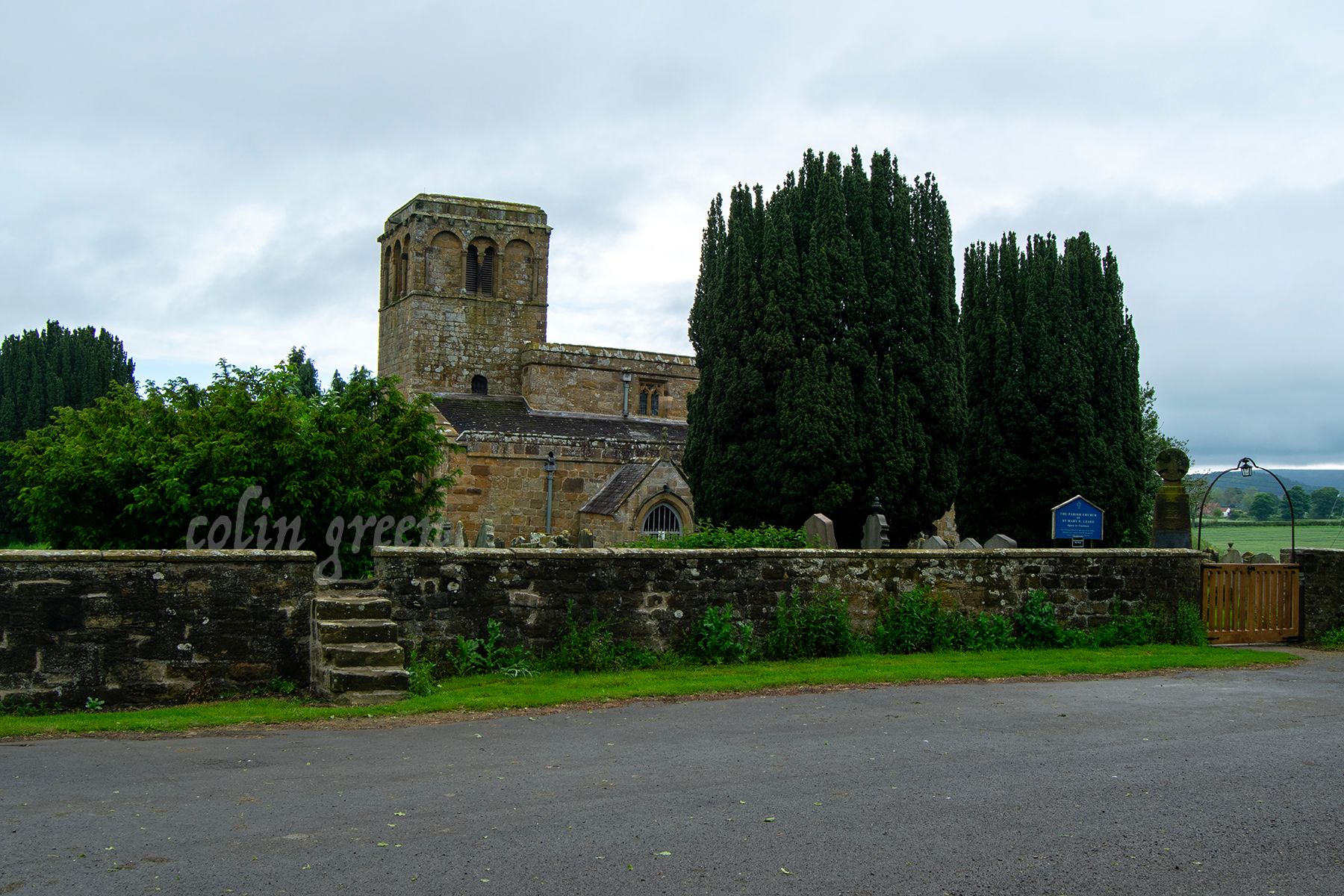 A church with a tall tower and a stone wall surrounding it. There are trees and a graveyard nearby. The sky is cloudy.