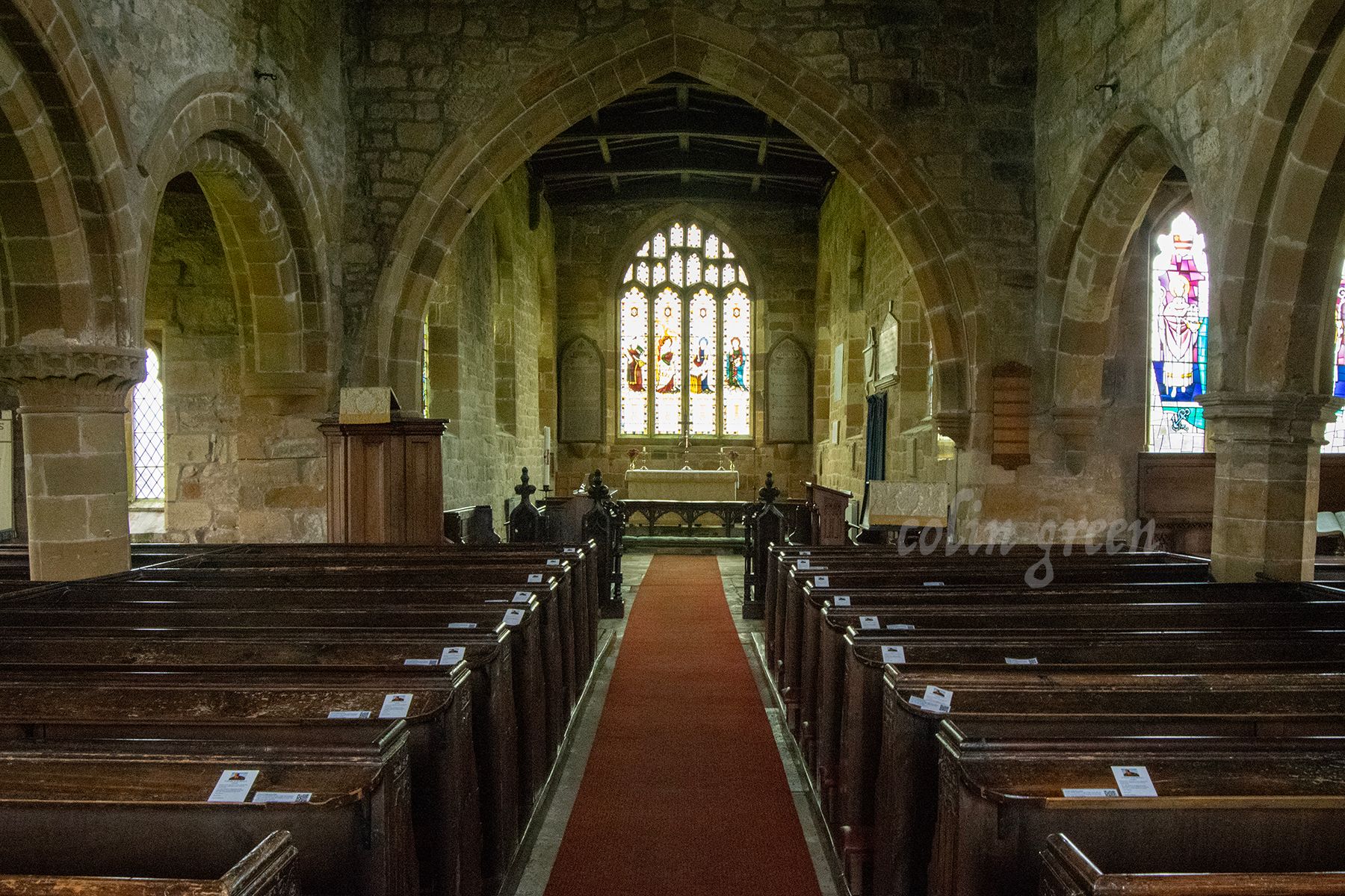 The interior of the Church of St Mary the Virgin, Leake with a stone archway, stained glass windows, and wooden pews. A red carpet runs down the center aisle.