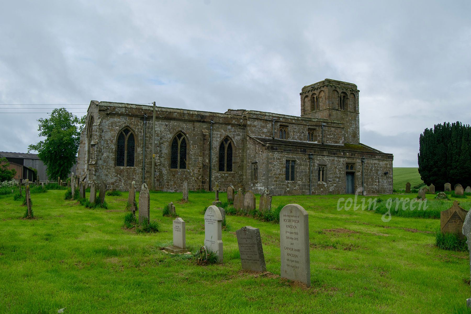 A church with a tall tower and a stone exterior, surrounded by a graveyard with tombstones. The sky is cloudy.