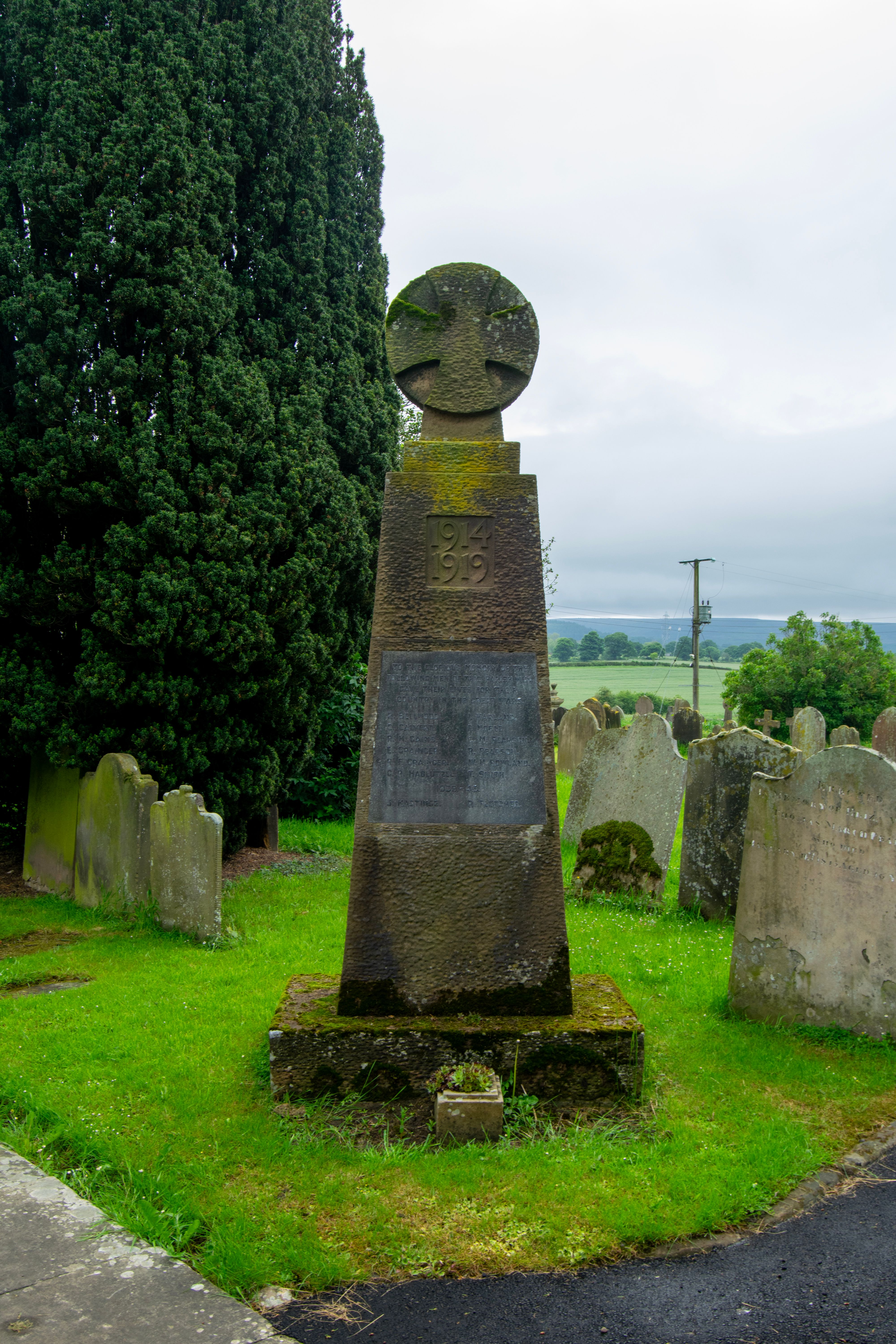 A stone war memorial in a graveyard. The memorial is topped with a cross and has a plaque with the inscription "GIGT 1914".