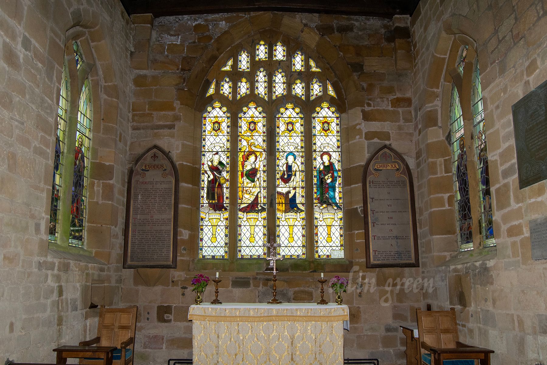 A stained glass window depicting religious figures, including Jesus and Mary, in a church. The window is surrounded by stone walls and illuminated by candles on a wooden altar.