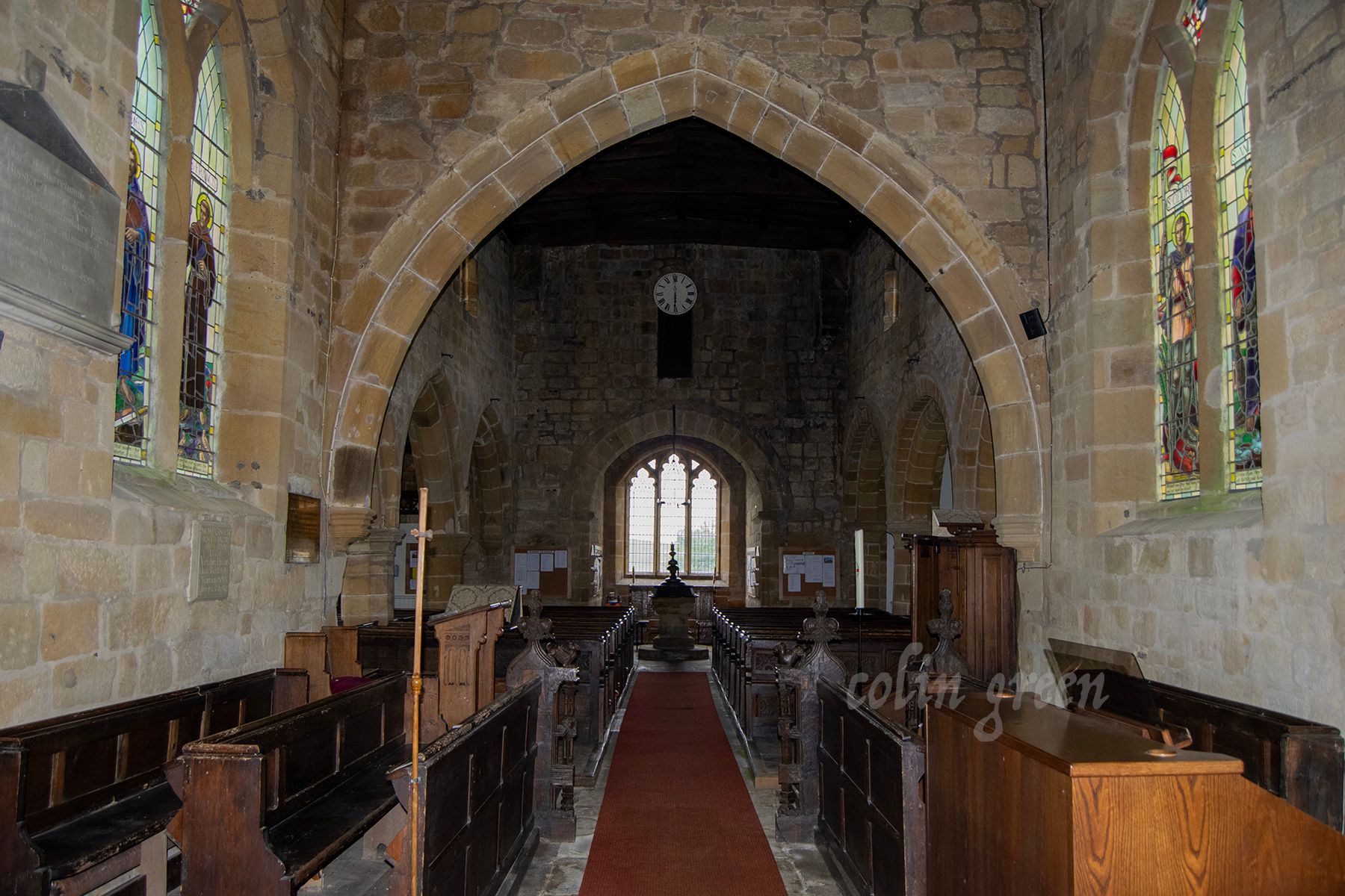 A long aisle in a stone church with a red carpet leading to the altar. Stained glass windows and wooden pews line the sides.