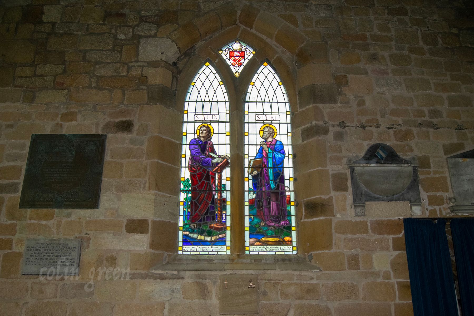 A stained glass window depicting two religious figures, likely apostles, standing on either side of a central figure. The window is set in a stone wall and surrounded by smaller windows.