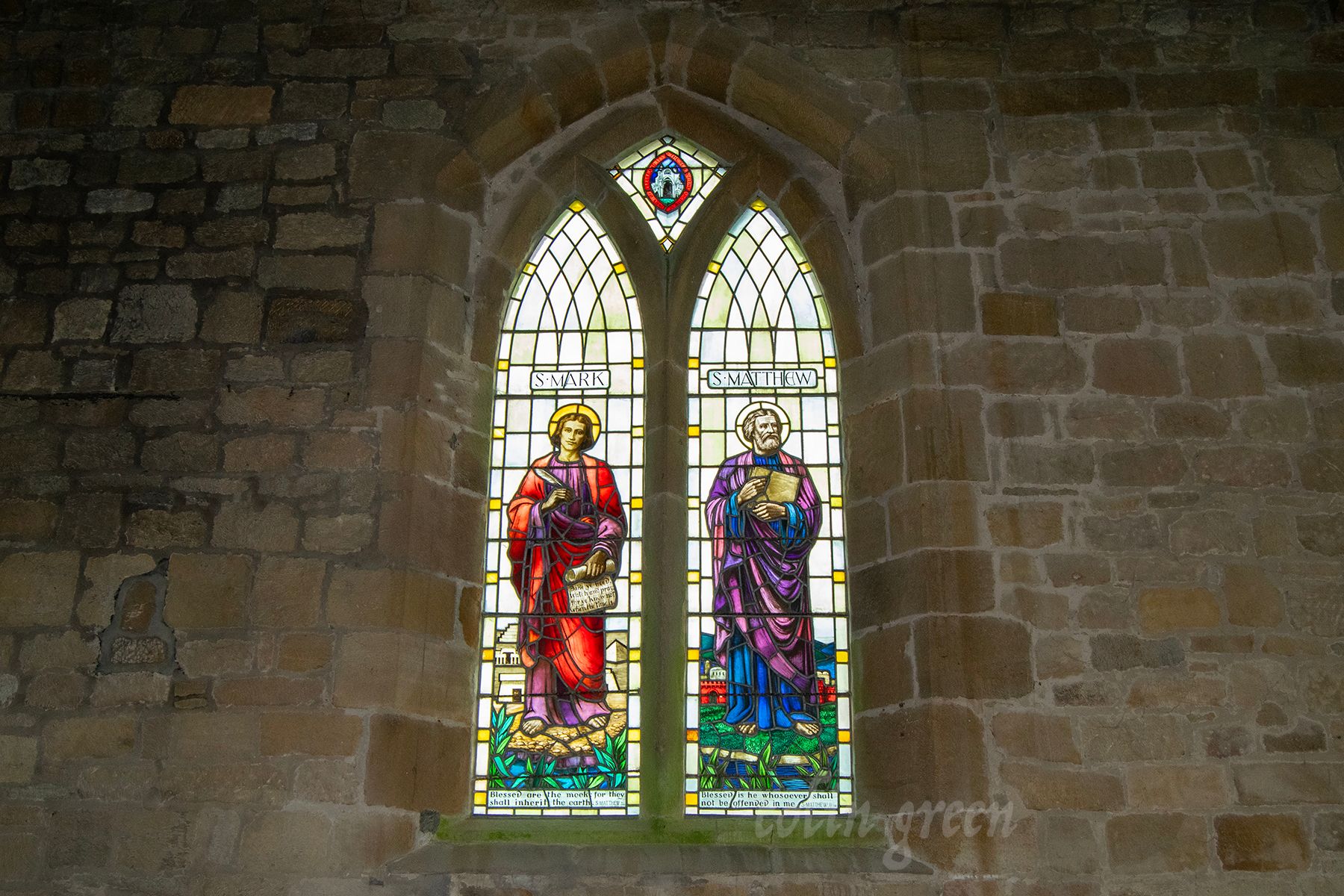 A stained glass window depicting St. Mark and St. Matthew, two of the Four Evangelists, standing side by side. The window is set in a stone wall and has a cross at the bottom.