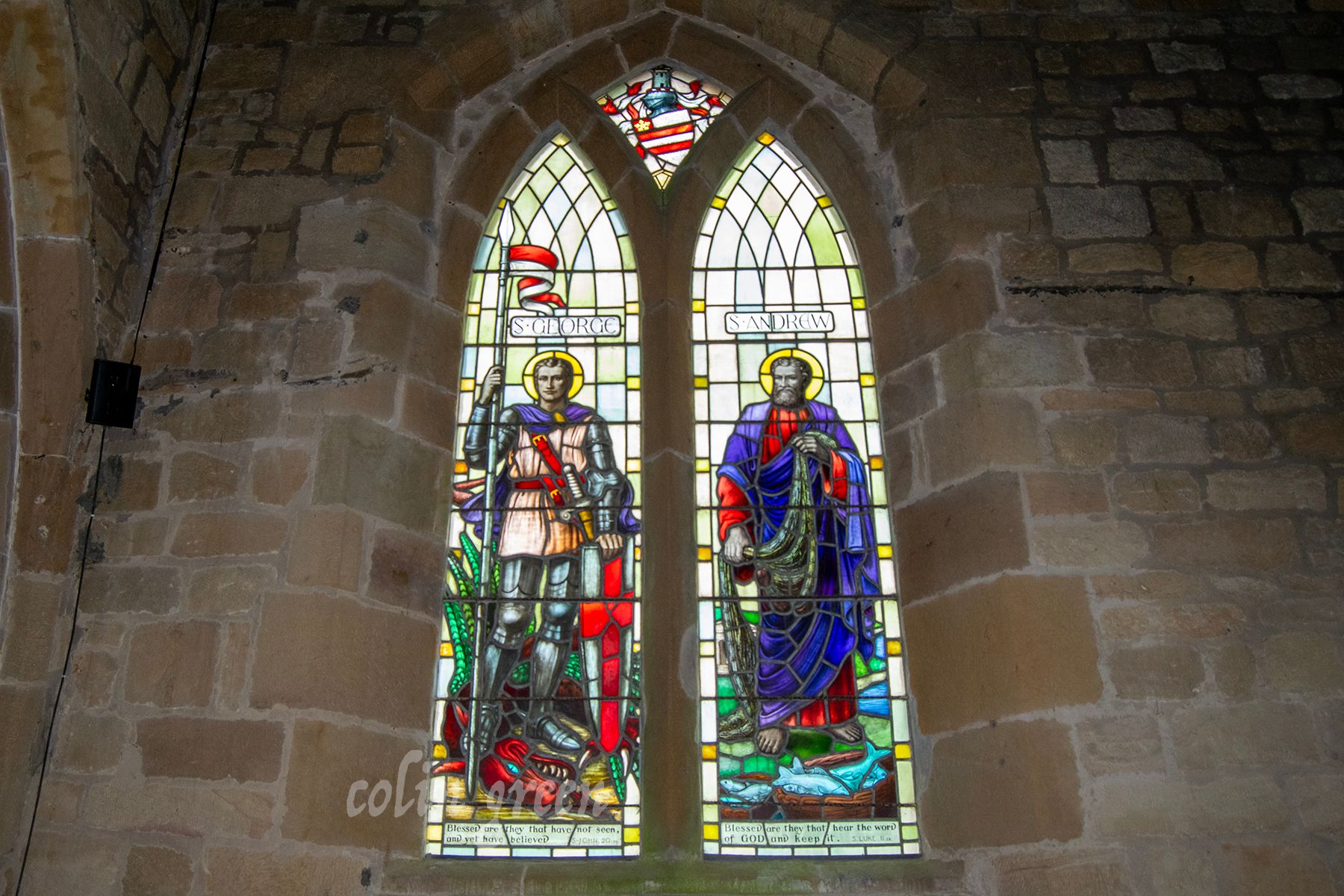 A stained glass window depicting St. George slaying a dragon on one side and St. Andrew, the patron saint of Scotland, on the other. The window is set in a stone wall.