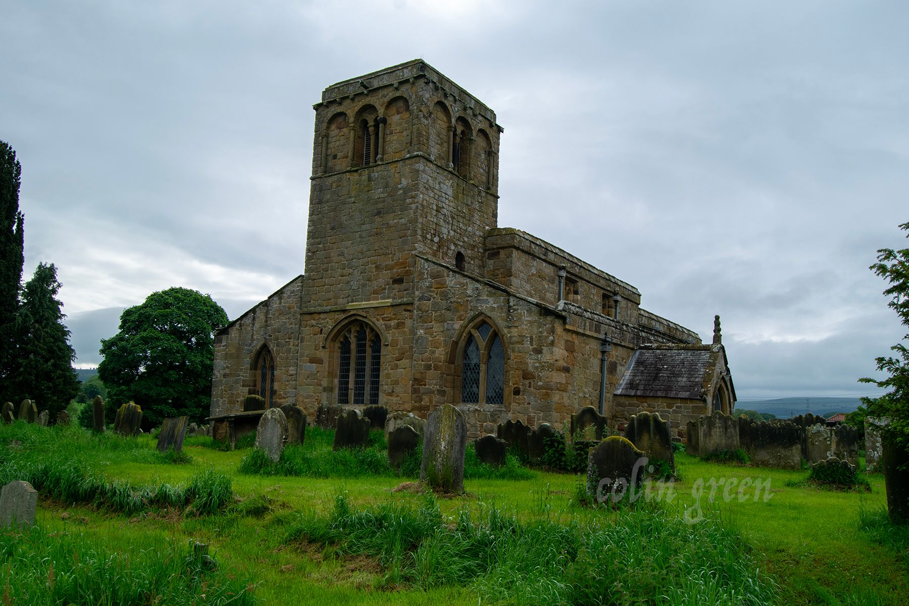 A historic stone church with a tall tower and a graveyard surrounding it. The church is surrounded by green fields and trees under a cloudy sky.