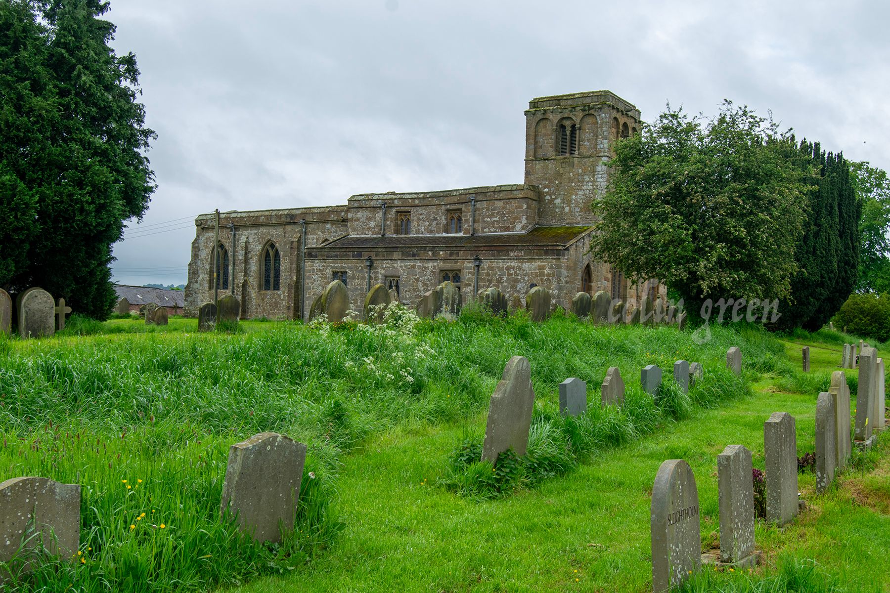 A historic stone church with a tall tower and a graveyard surrounding it. The church is surrounded by green fields and trees under a cloudy sky.