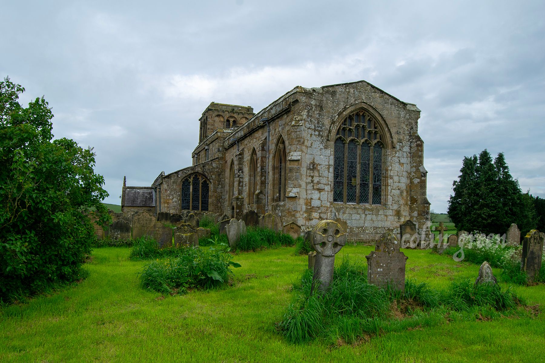 A historic stone church with a tall tower and a graveyard surrounding it. The church is surrounded by green fields and trees under a cloudy sky.