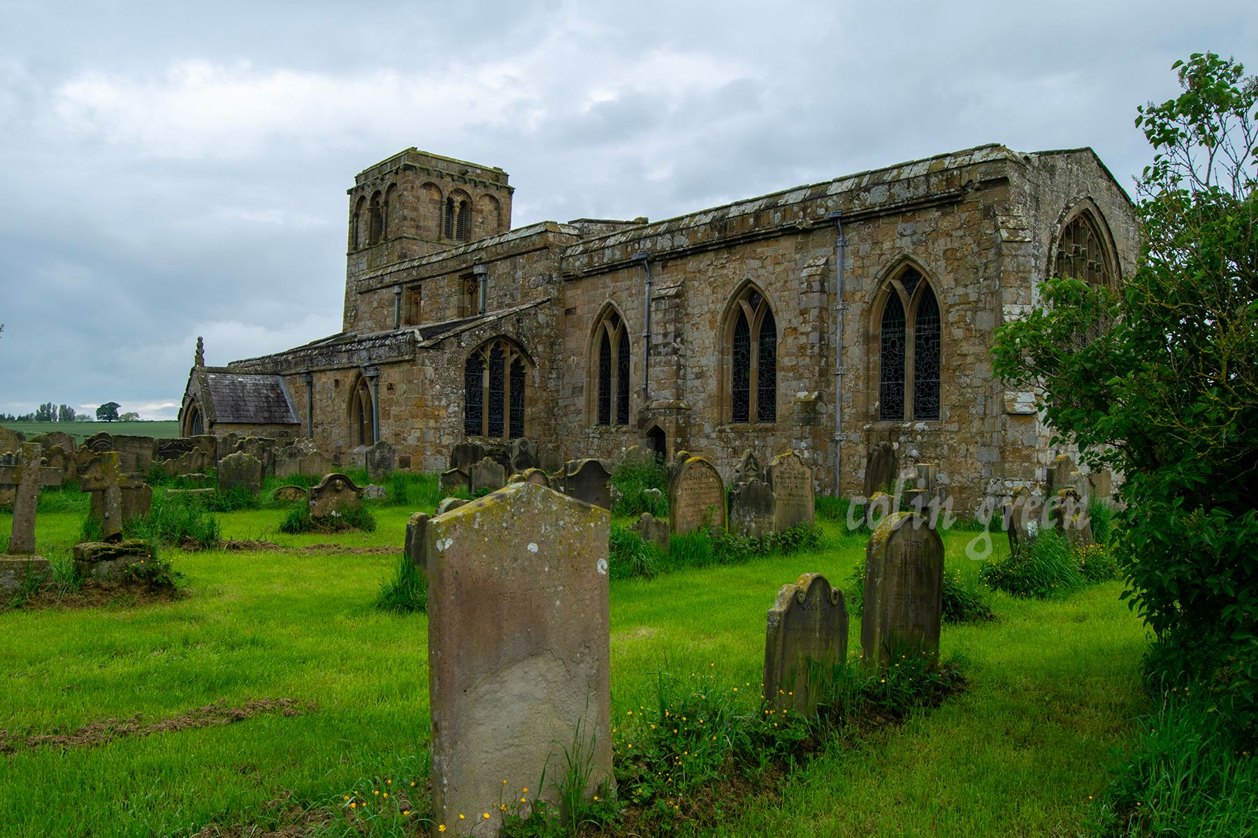 A historic stone church with a tall tower and a graveyard surrounding it. The church is surrounded by green fields and trees under a cloudy sky.