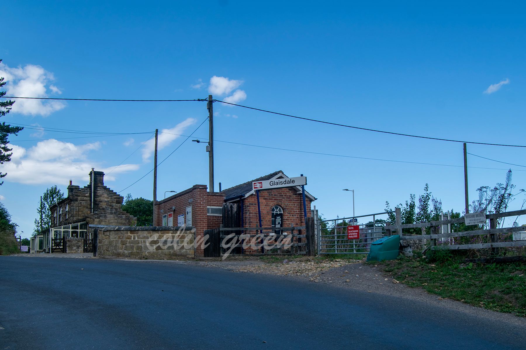 Glaisdale Railway Station with a brick building entrance and a sign reading "Glaisdale." The building is surrounded by a fence. A road leads up to the station, with a clear blue sky above.