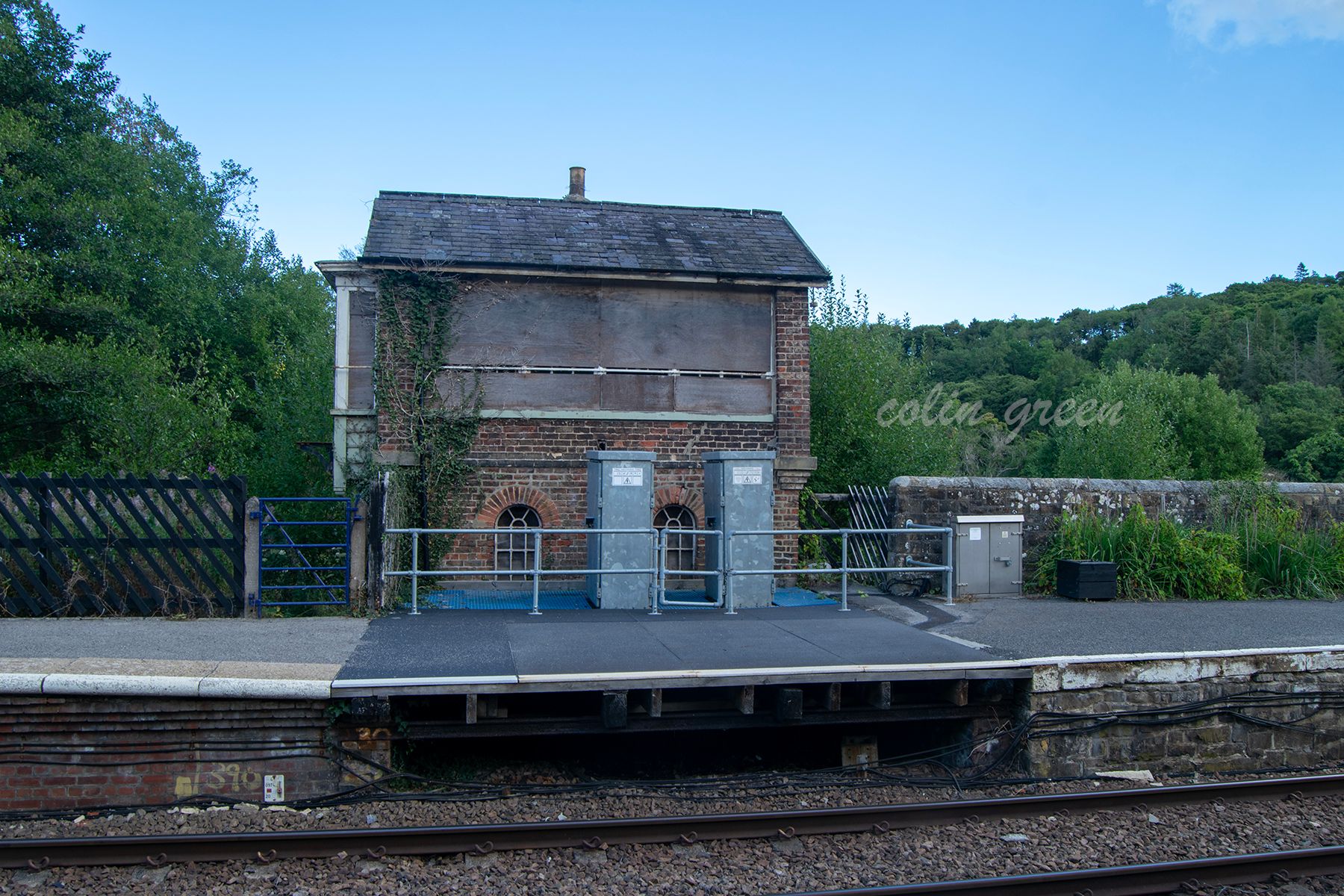 A disused signal box with a brick exterior and a slate roof. The building is partially covered in ivy and has a boarded-up window. The signal box stands on a platform next to a railway track.