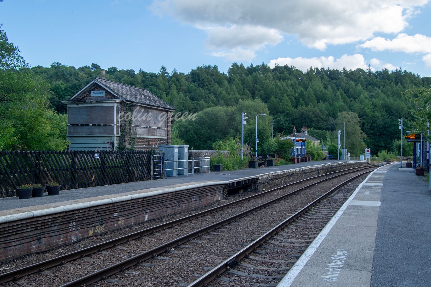 A view of Glaisdale Railway Station, showing the platform, tracks, and a disused signal box. The station is surrounded by lush green hills and trees.