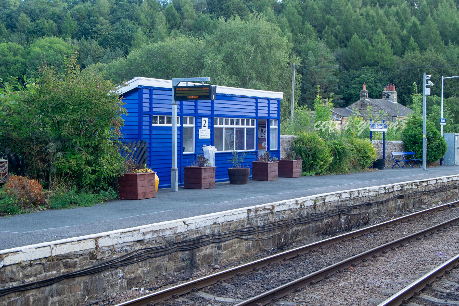 A small, blue-painted station building with a waiting room and platform. The station is surrounded by greenery and overlooks a railway track.