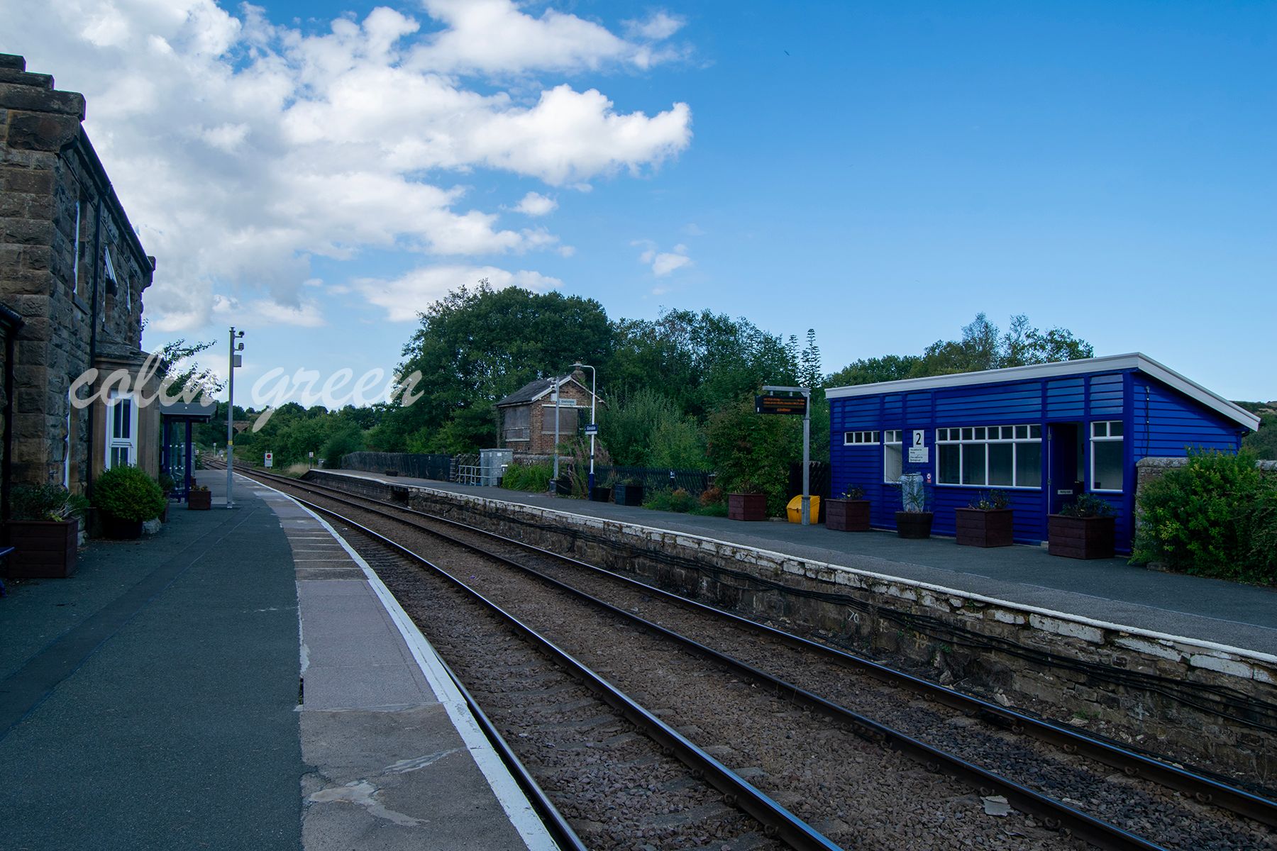 A view of Glaisdale Railway Station, showing the platform, tracks, and a disused signal box. The station is surrounded by lush green hills and trees.