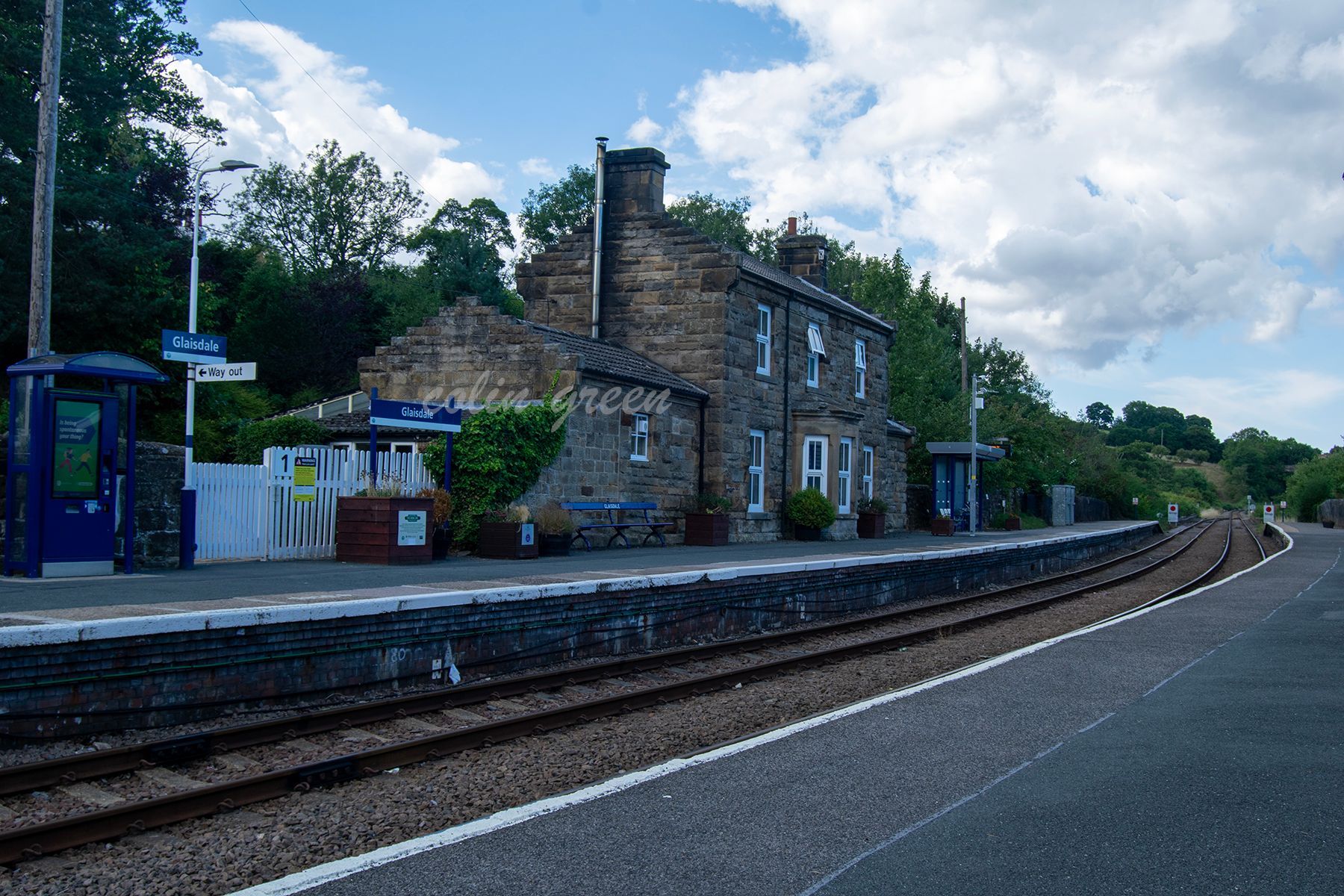 A view of Glaisdale Railway Station, showing the platform, tracks, and a disused signal box. The station is surrounded by lush green hills and trees.