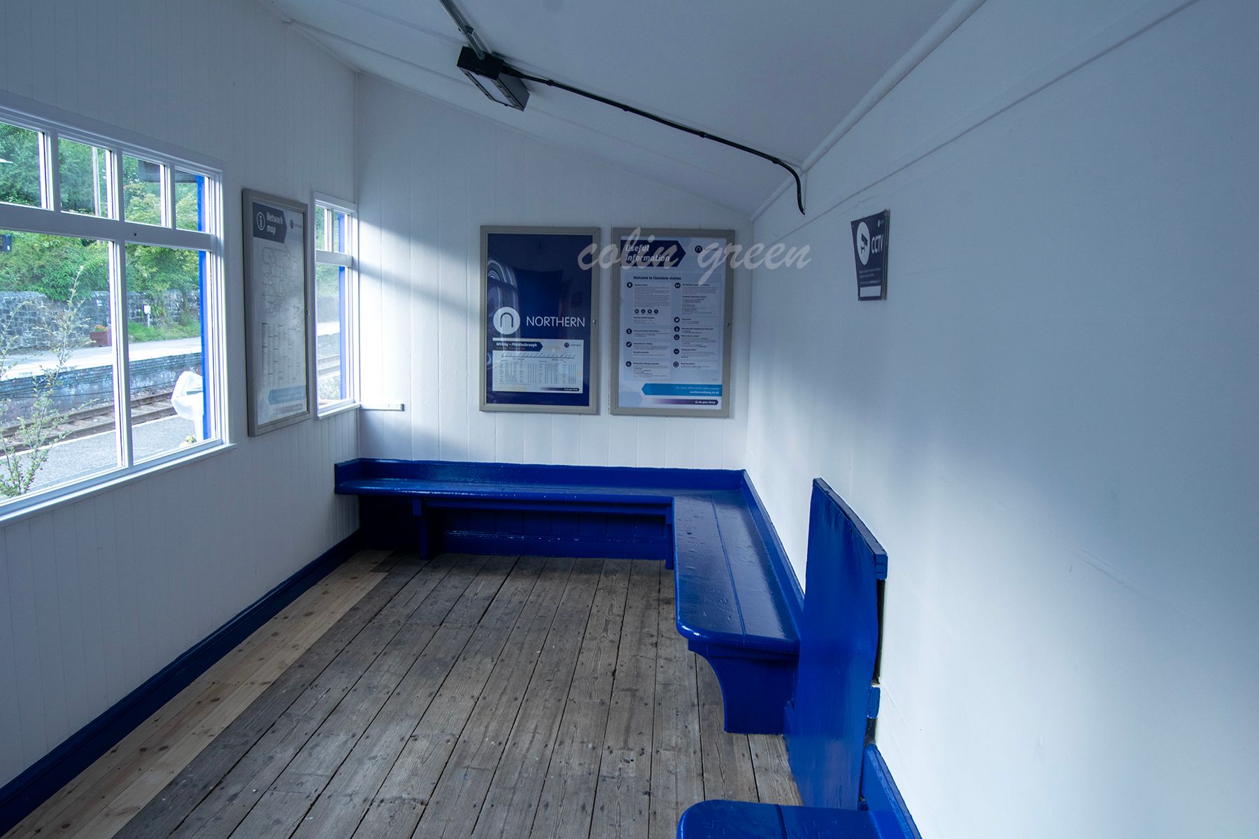 The interior of a small, white-walled waiting room at Glaisdale Railway Station. The room features wooden floors, blue benches, and a Northern Rail poster.