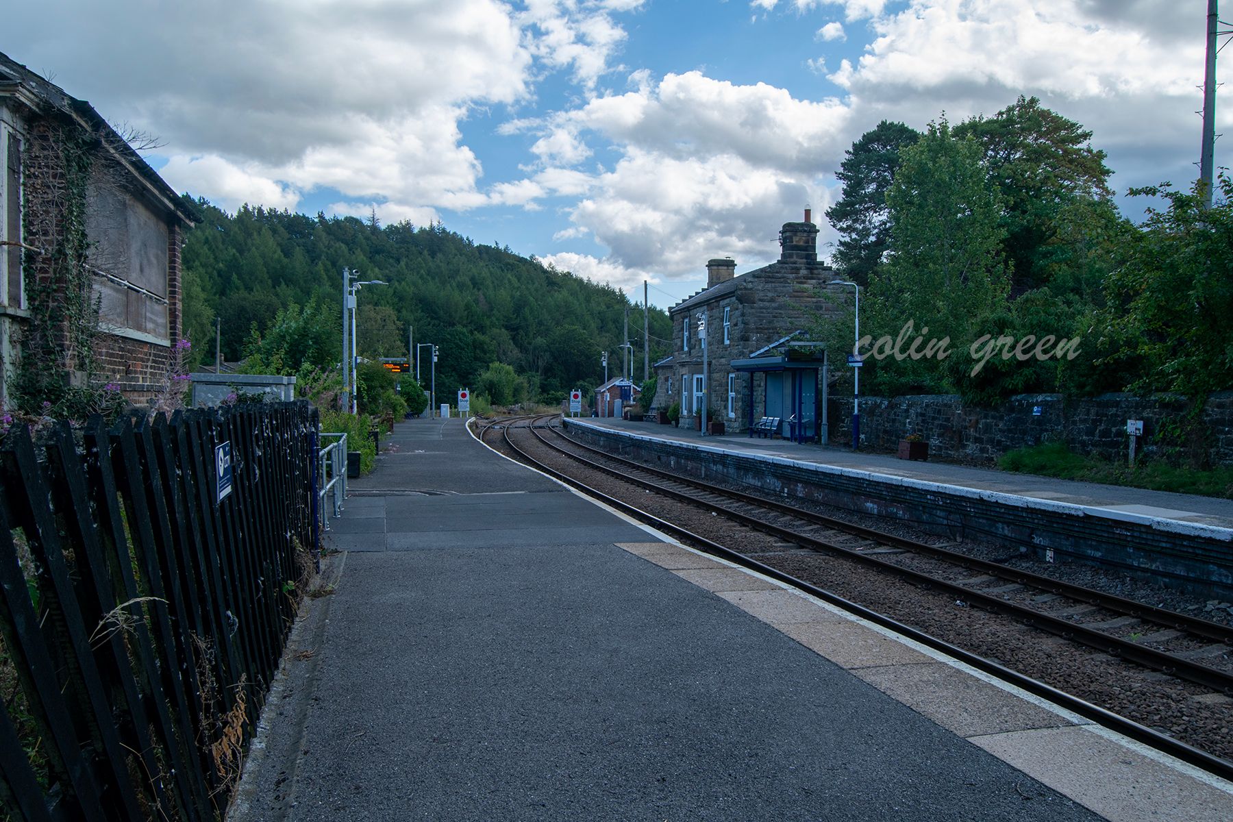 A view of Glaisdale Railway Station, showing the platform, tracks, and station buildings. The station is surrounded by lush green hills and trees.