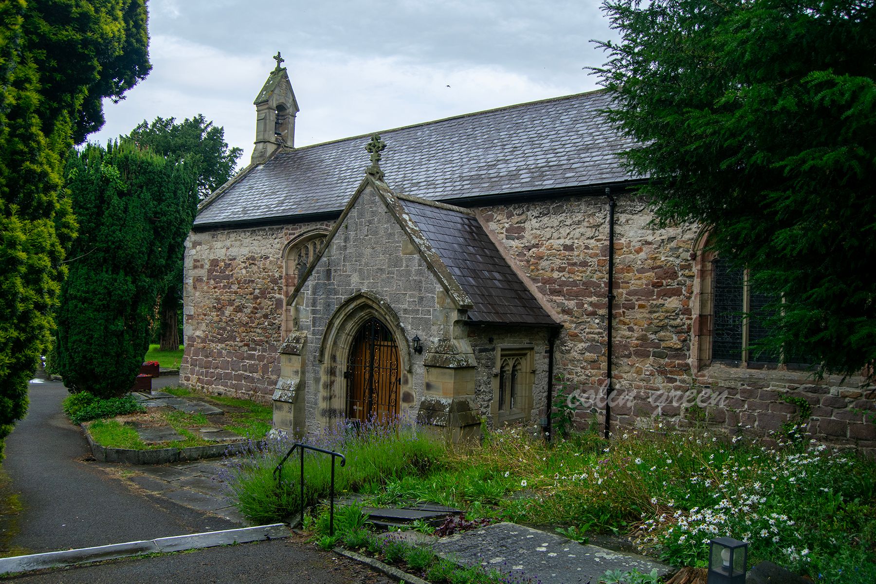 A stone church with a slate roof and a prominent arched doorway. There is a small steeple with a cross on top. The church is surrounded by trees and a grassy area with wildflowers.