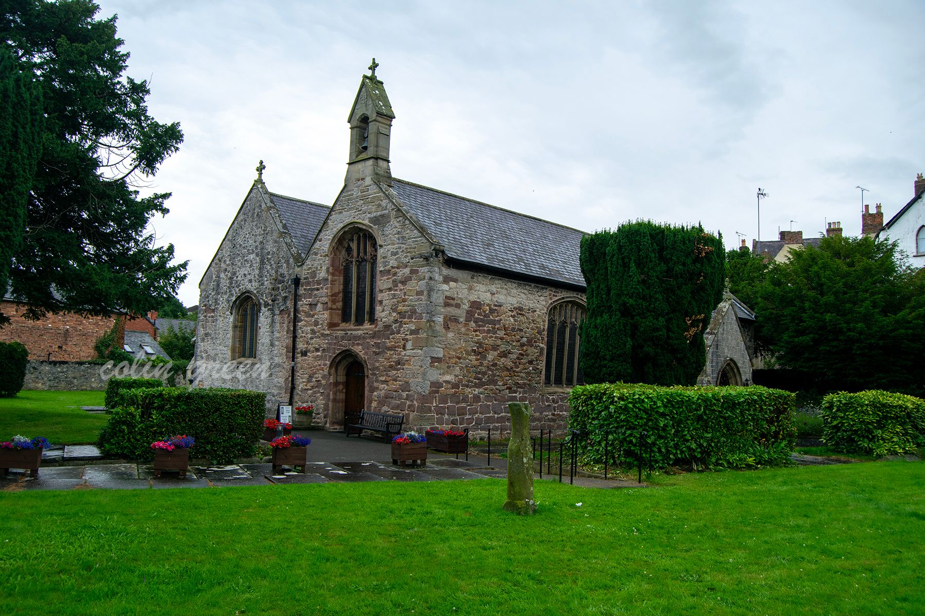 A stone church with a slate roof and a prominent arched doorway. There is a small steeple with a cross on top. The church is surrounded by trees, a grassy area, and flowerpots.