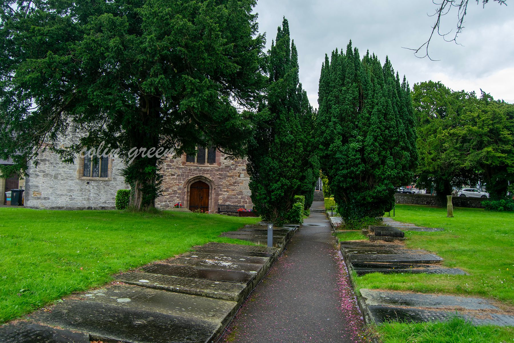 A stone church with a slate roof and a prominent arched doorway. The church is surrounded by trees, a grassy area, and a pathway leading to a graveyard with gravestones.