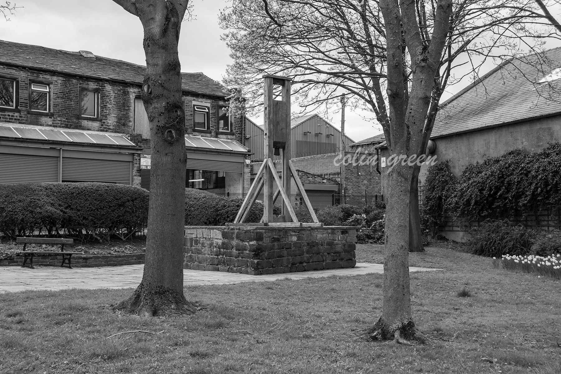 A black and white photo of a guillotine in a small park, surrounded by trees and buildings.