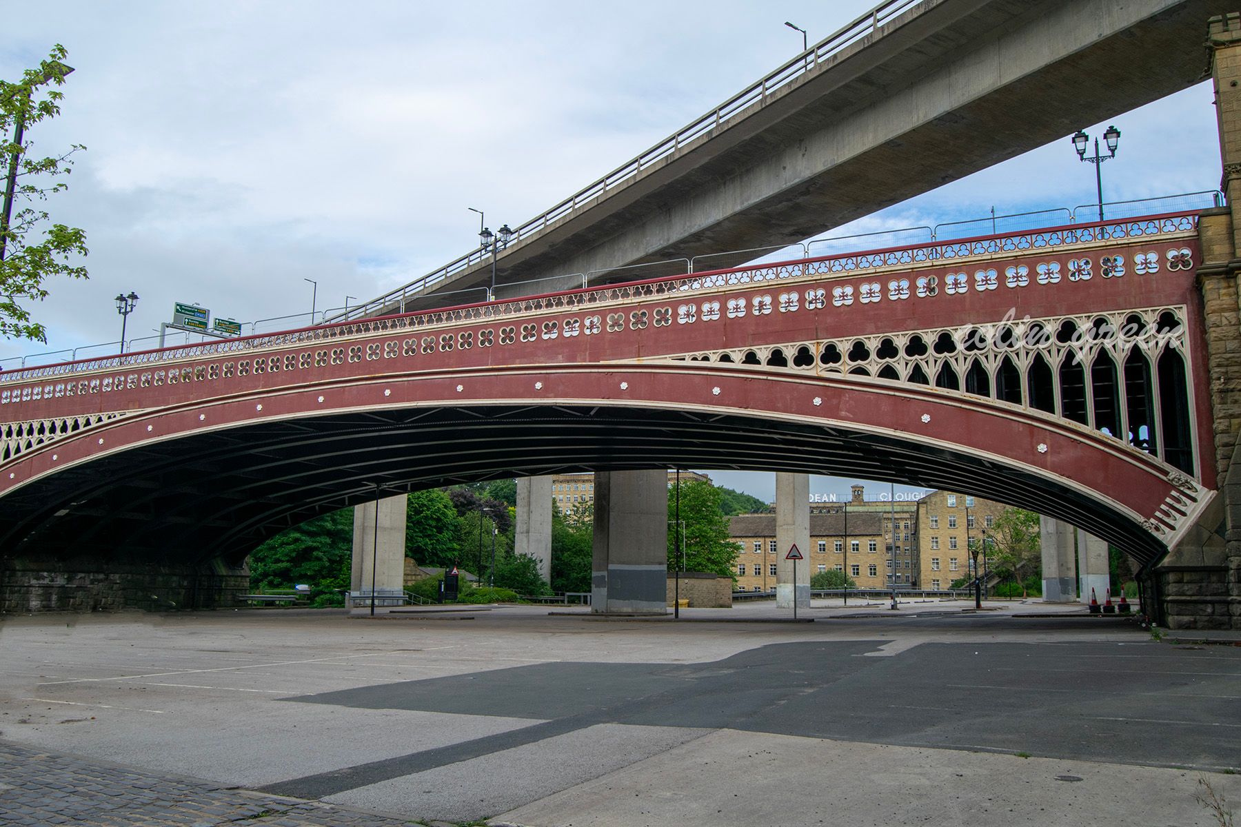 A view of the North Bridge in Halifax, England, with its distinctive red arches and intricate detailing. A modern road bridge spans above it