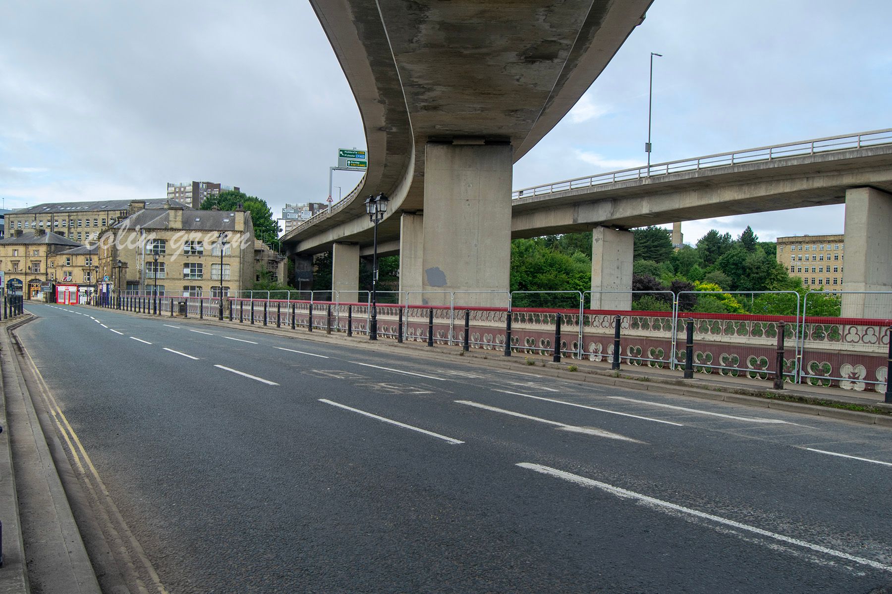 A view of the North Bridge in Halifax, England, from below. The modern road bridge arches overhead, creating a dramatic contrast with the quiet street below.