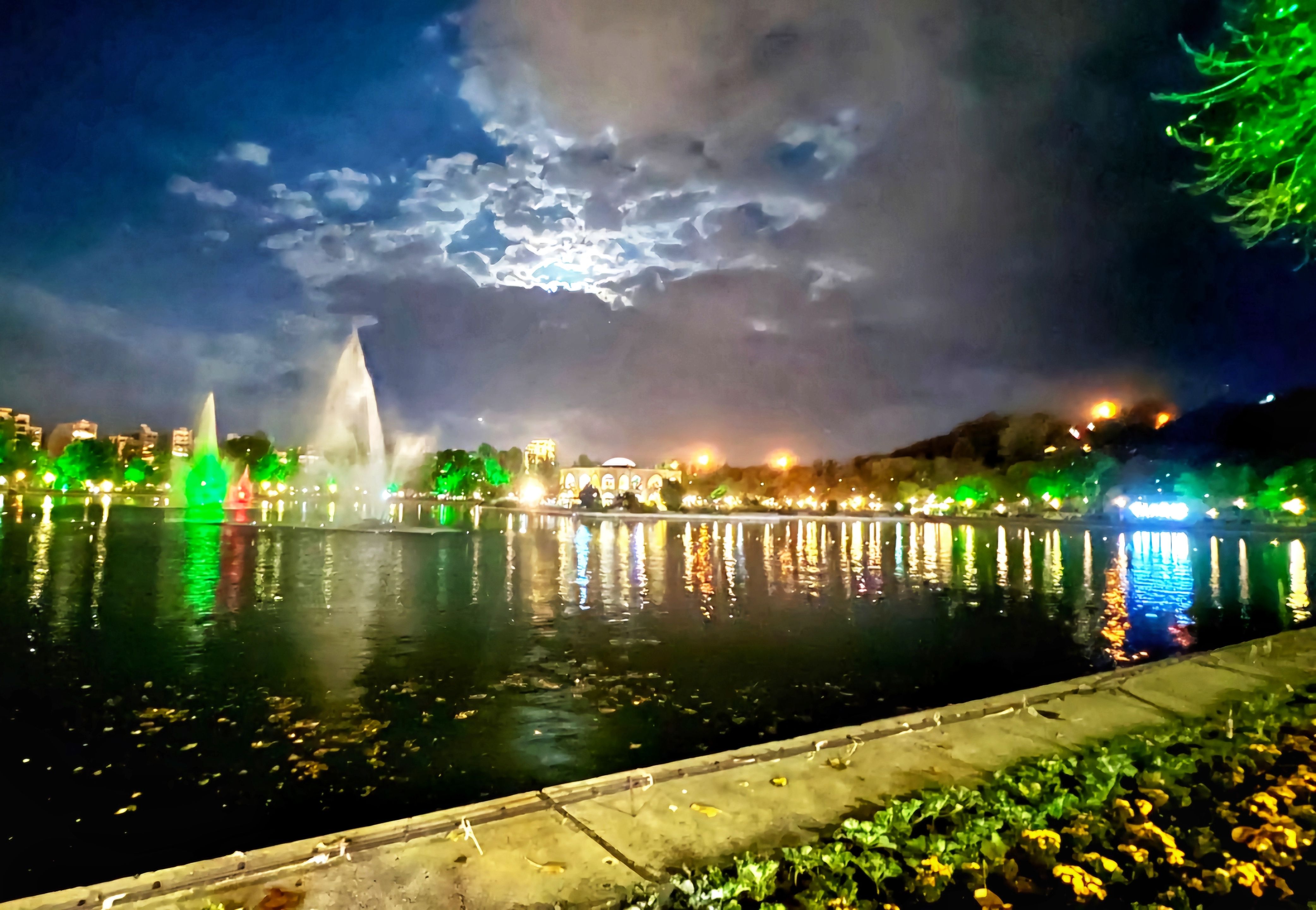 A nighttime scene in a park with lights softly illuminating the surroundings. The calm waters of a pond reflect the lights, creating a peaceful and dreamy atmosphere.