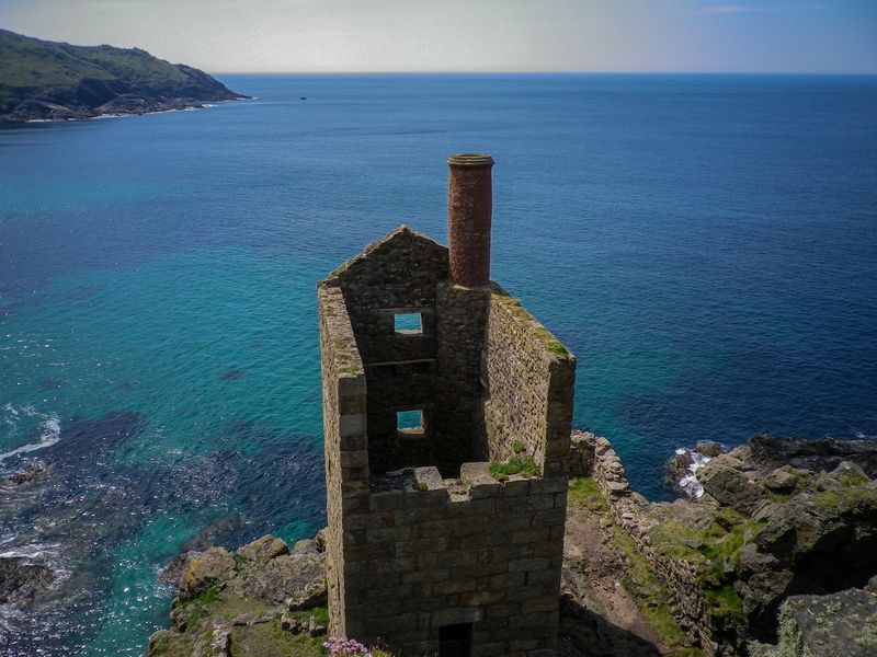 A ruined tin mine engine house stands sentinel like on cliffs overlooking a turquoise blue ocean.