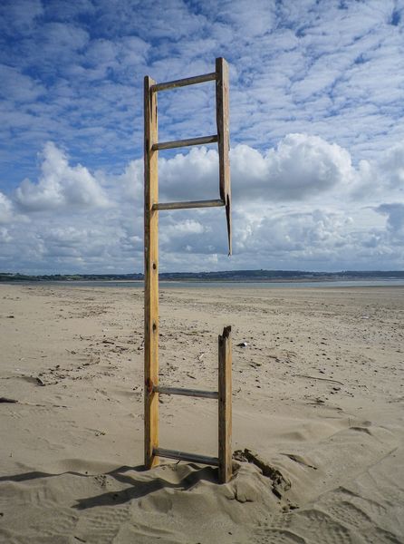 An unsupported broken rungs step ladder reaches toward a blue sky with fluffy white clouds from a sandy beach