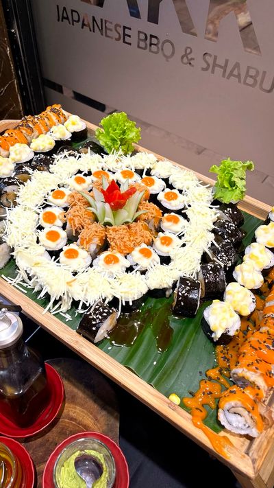 A beautifully arranged sushi platter sits on a wooden table, showcasing an elegant assortment of colors and textures. The platter features a variety of sushi rolls, including vibrant slices of sashimi, delicate nigiri, and carefully crafted maki rolls with fresh fish, avocado, and cucumber. The sushi is artistically arranged with some pieces garnished with a sprinkle of sesame seeds, thin slices of radish, and a hint of wasabi on the side. Small dishes of soy sauce and pickled ginger are placed alongside, completing the aesthetic. The entire setup exudes a sense of freshness and sophistication, ready to be enjoyed in a delightful culinary experience.